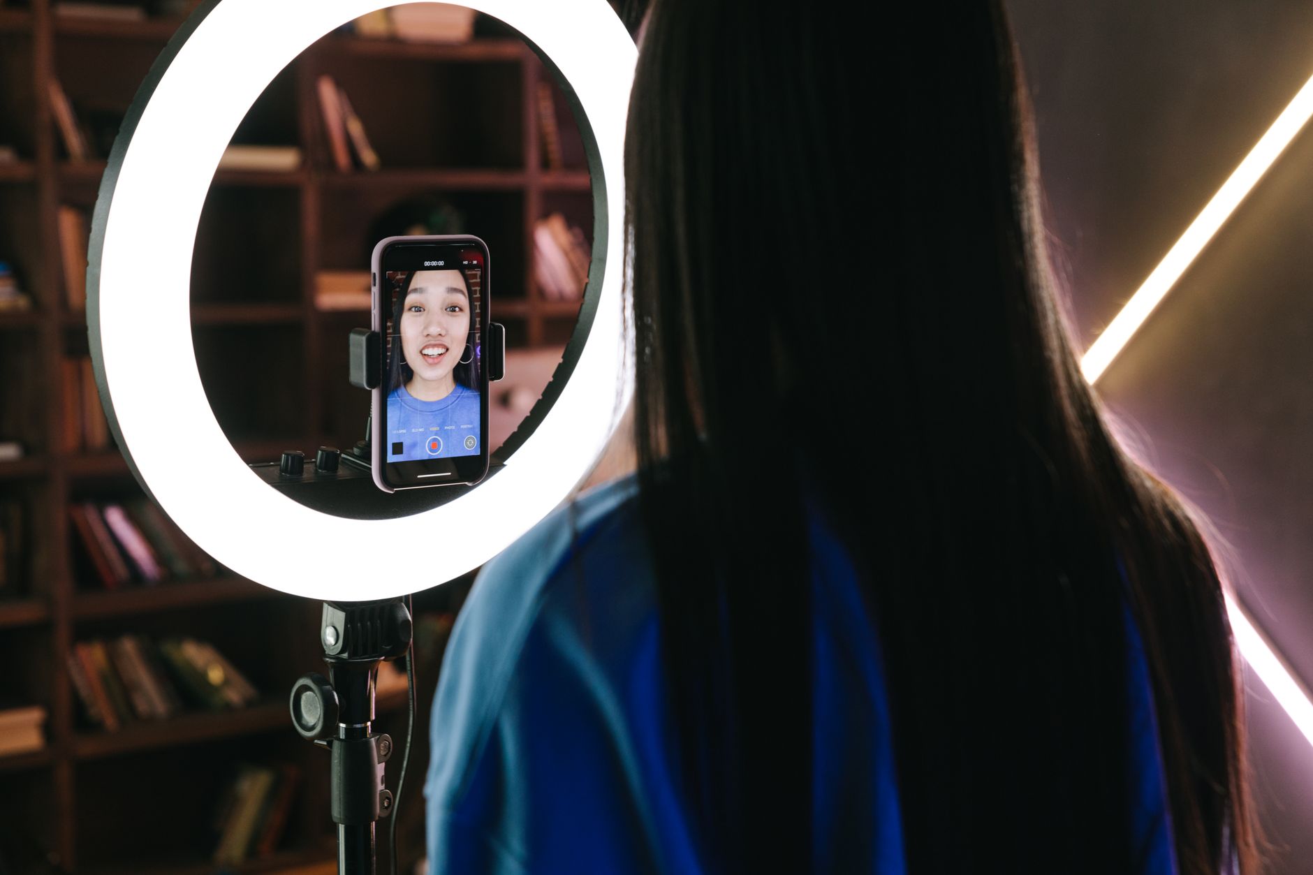 Woman seen from behind facing a ring light with a smartphone on a stand, the phone screen showing her smiling as she records a video